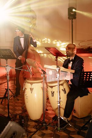 Two musicians in tuxedos perform jazz, one playing double bass and another at vibraphone with conga drums on a warmly lit stage.