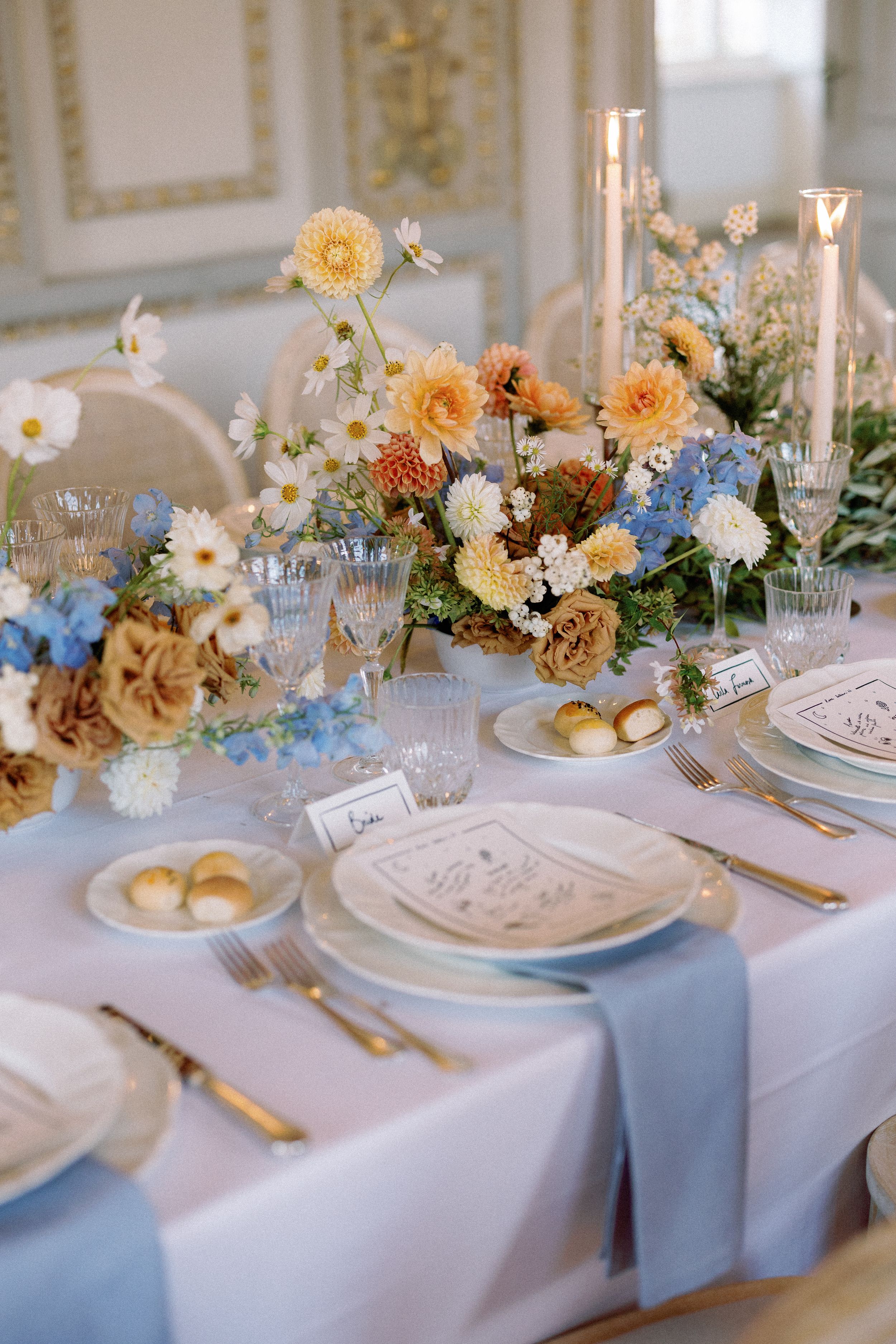 Elegant wedding reception table with blue runner, gold cutlery, floral centerpieces in blue, orange, and white, and crystal glassware.