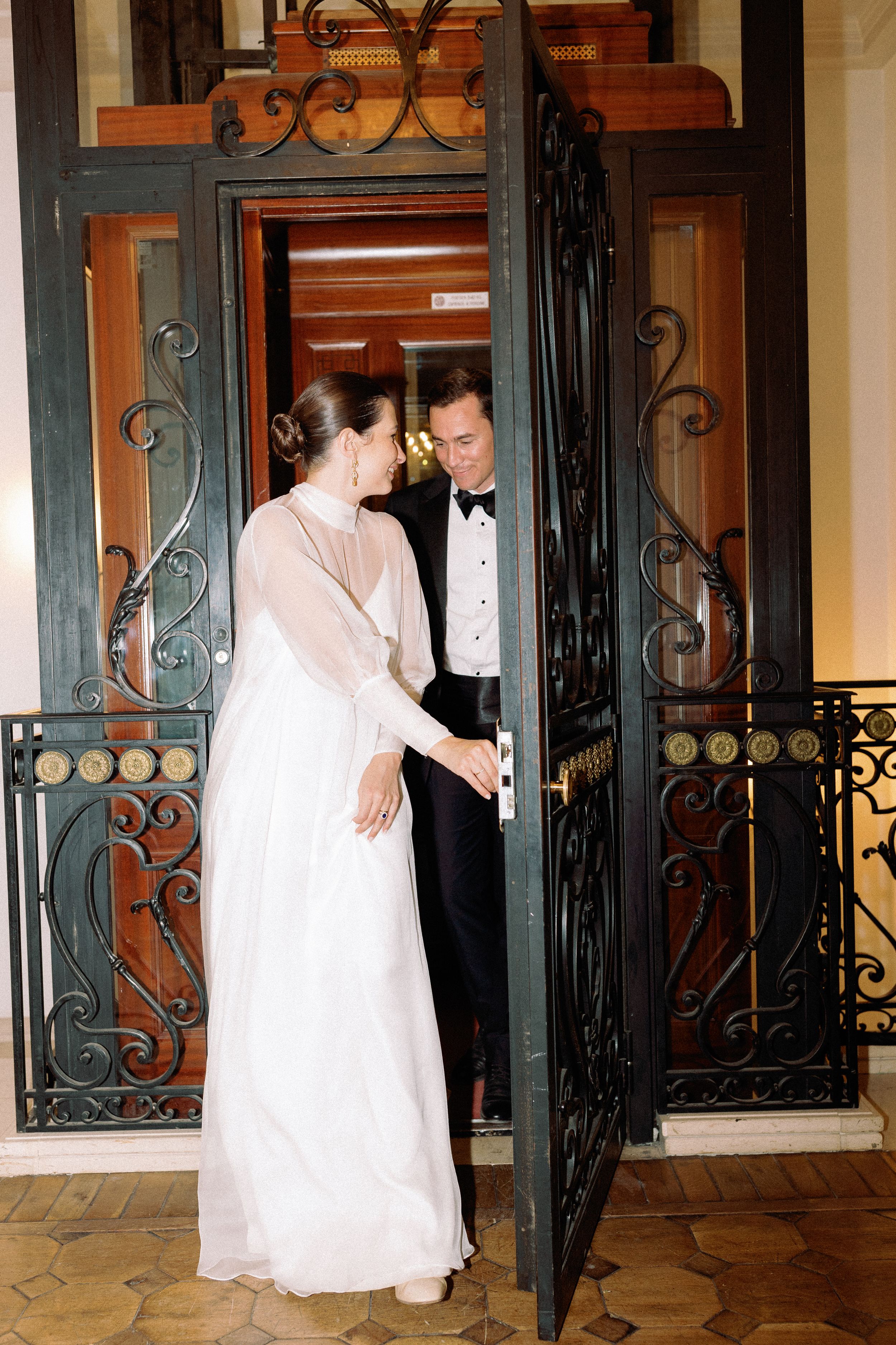 Elegant bride and groom holding hands in a doorway with ornate black iron doors at their wedding venue.