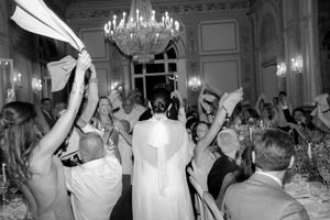 Black and white photo of a bride entering a grand ballroom as wedding guests wave napkins in celebration.