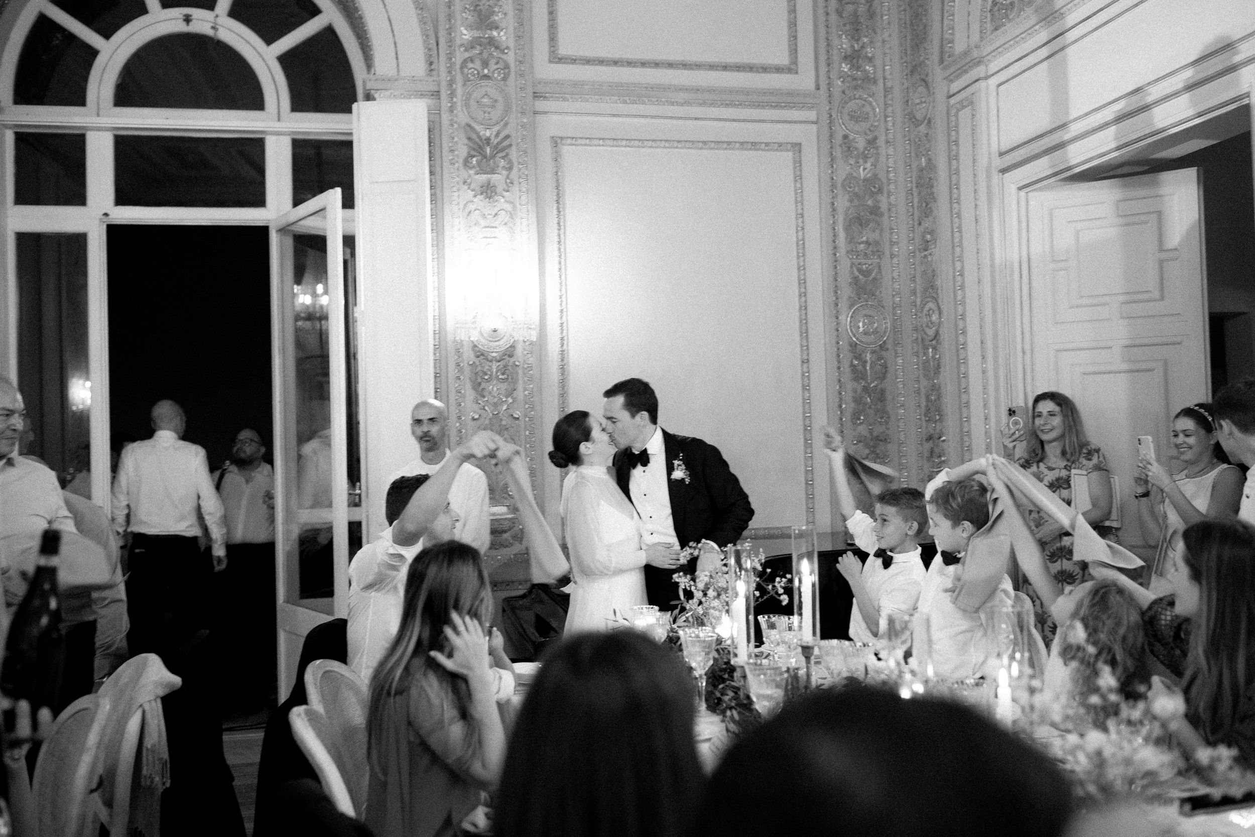 Bride and groom share a kiss during their elegant wedding reception as guests celebrate.