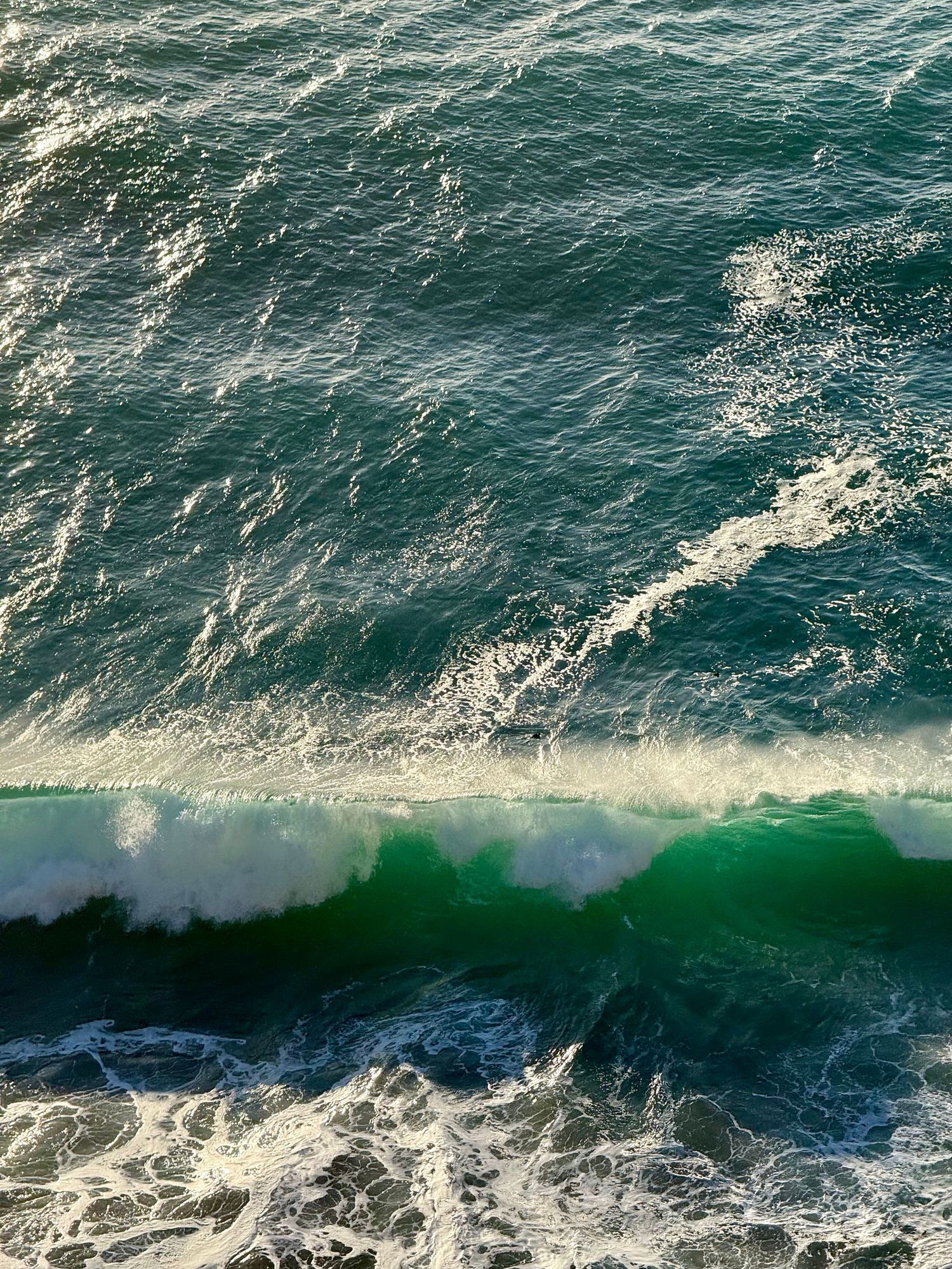 Aerial view of vibrant turquoise ocean wave crashing with white sea foam in sunlit water.