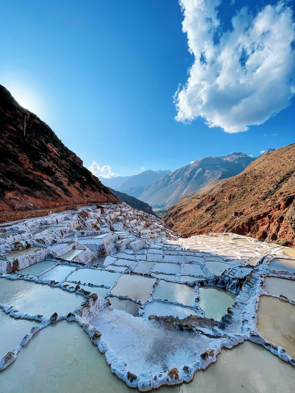 Ancient Maras salt ponds terraced into Andean mountainside with snow-white salt flats under blue sky.