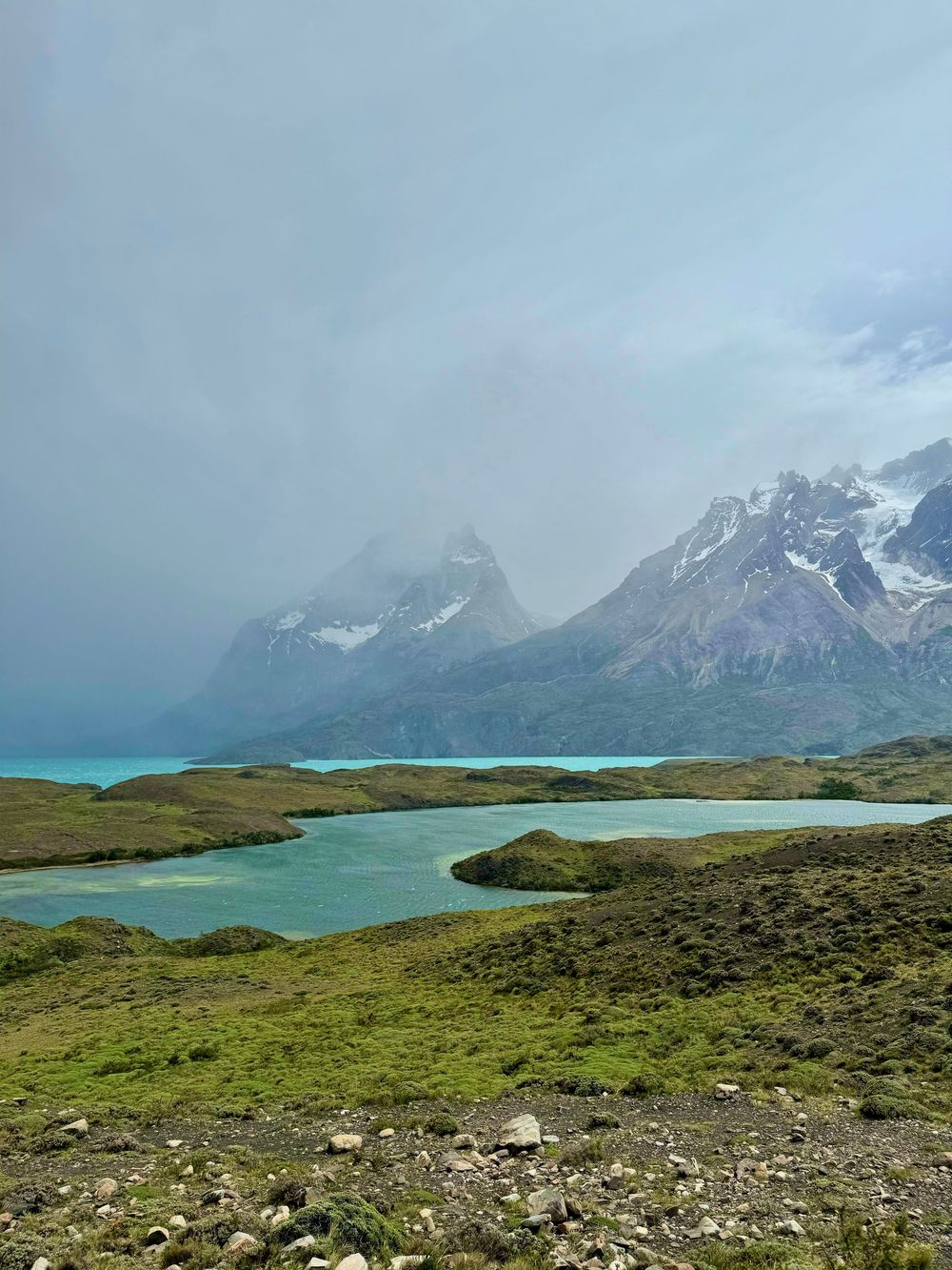 Turquoise glacial lakes wind through lush green landscape below misty snow-capped Patagonian mountain peaks.