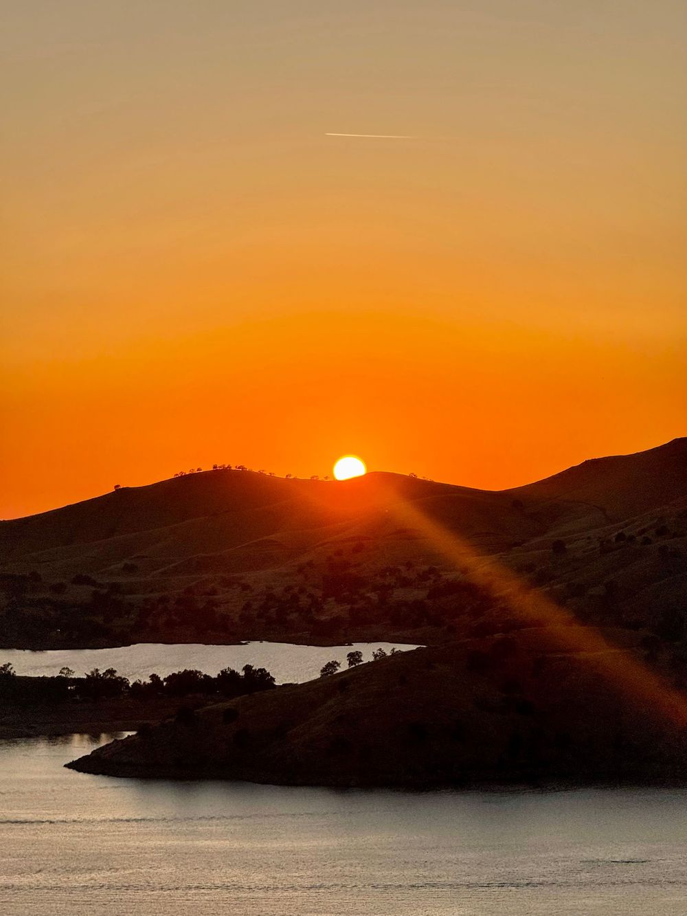 A brilliant orange sunset sinks behind silhouetted hills reflecting golden light over a calm lake.