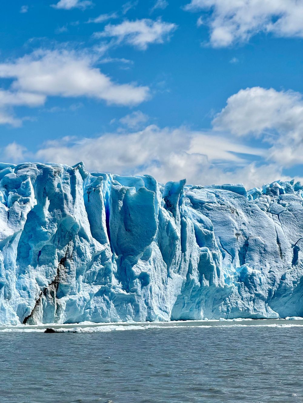 Massive blue ice wall of Perito Moreno Glacier rises from dark water under a bright blue sky.
