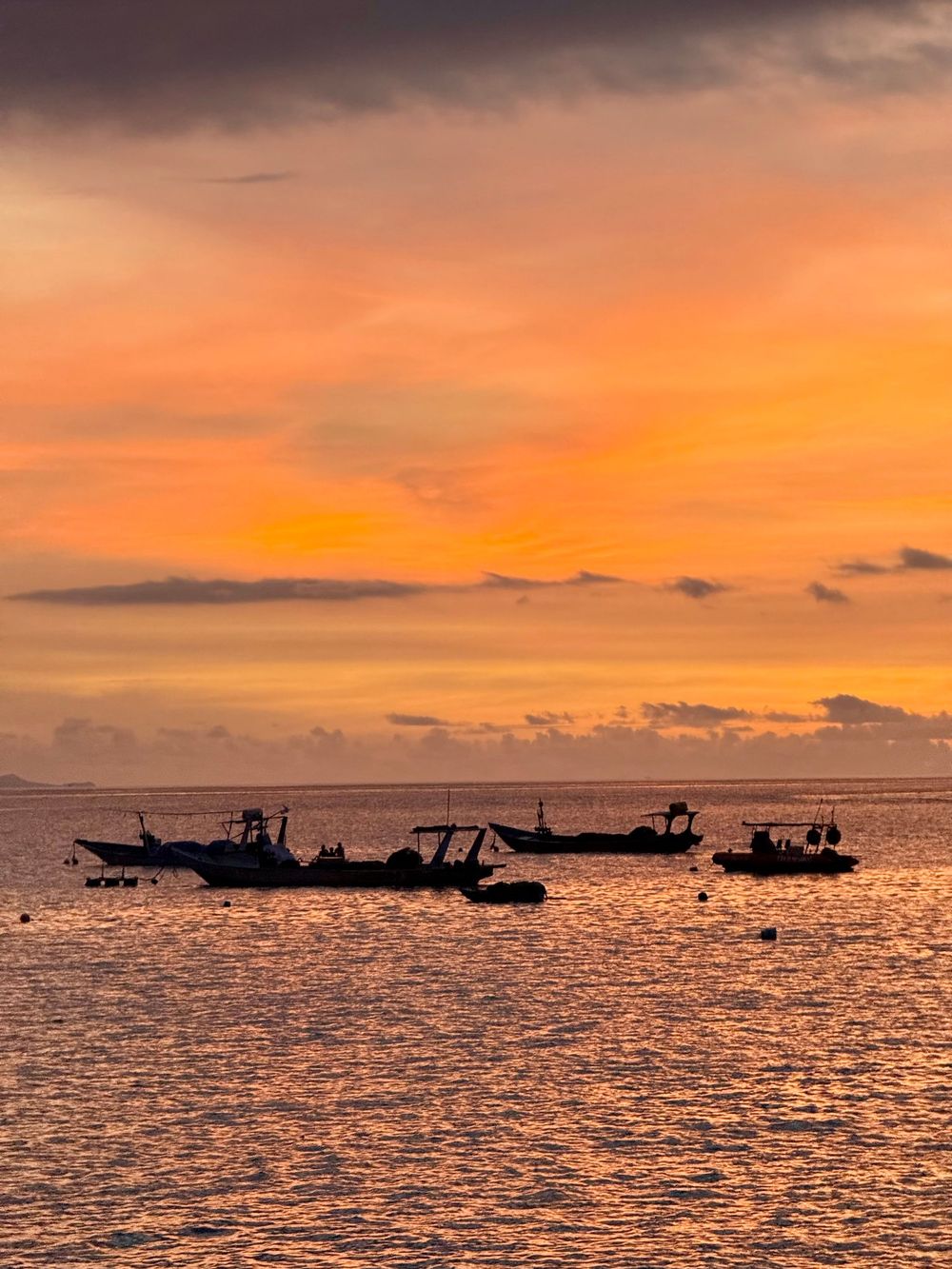 Silhouetted boats anchor on calm ocean waters beneath a dramatic orange and gold tropical sunset sky in Lombok.