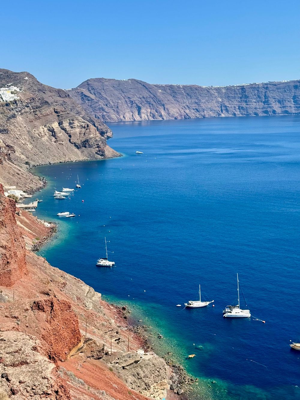 Aerial view of white sailboats anchored in vivid blue Santorini caldera waters with dramatic volcanic cliffs.