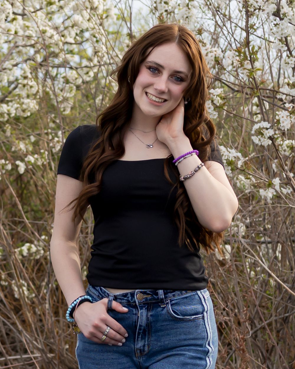 Young woman with long auburn hair smiling outdoors, wearing black crop top and jeans, with white flowering trees in background.