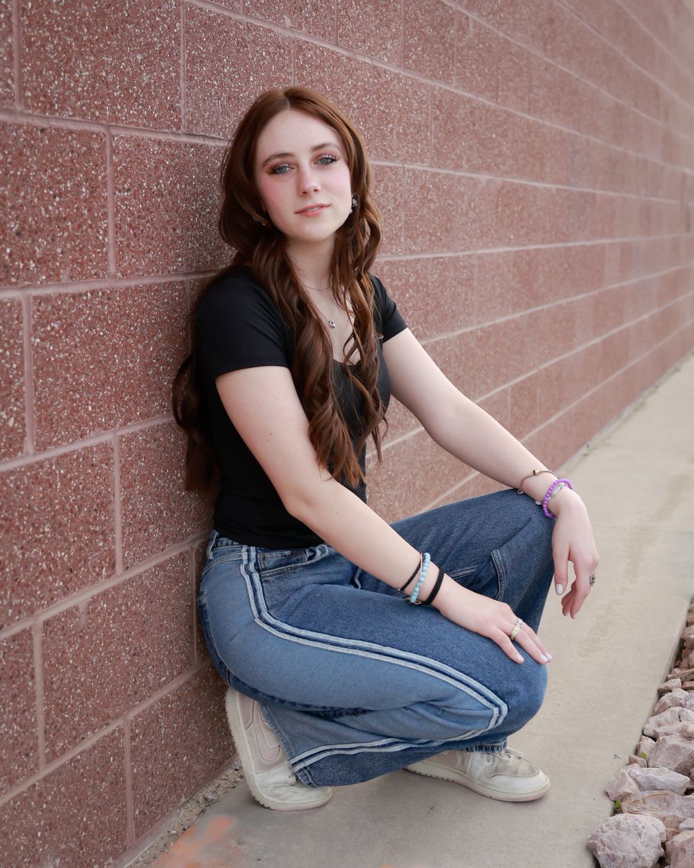 A young woman with long red hair in jeans and black top poses against a brick wall outdoors.