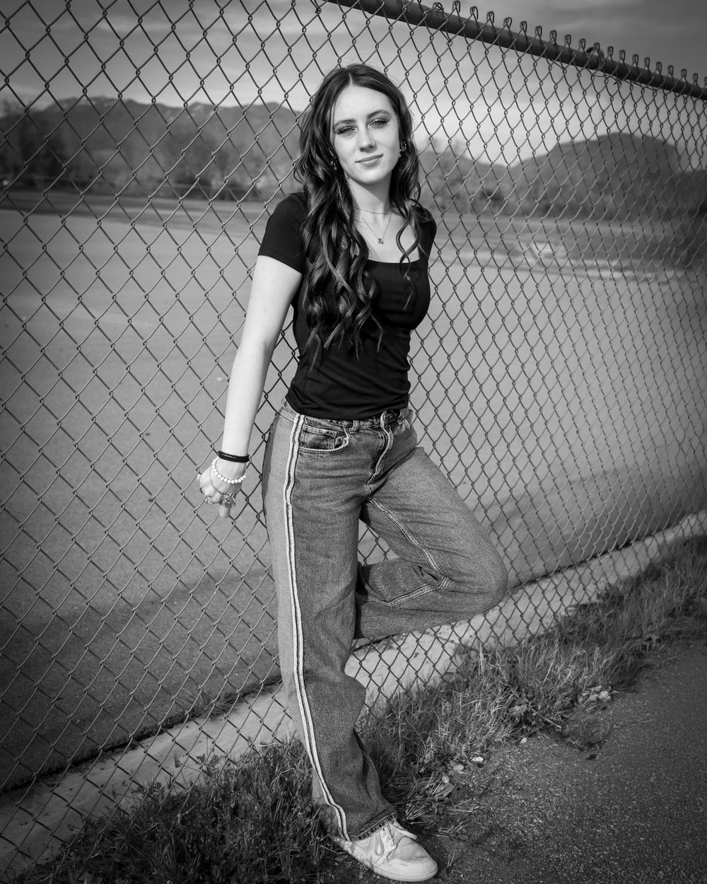 Young woman in black top and striped jeans posing against a chain-link fence in a black and white photo.