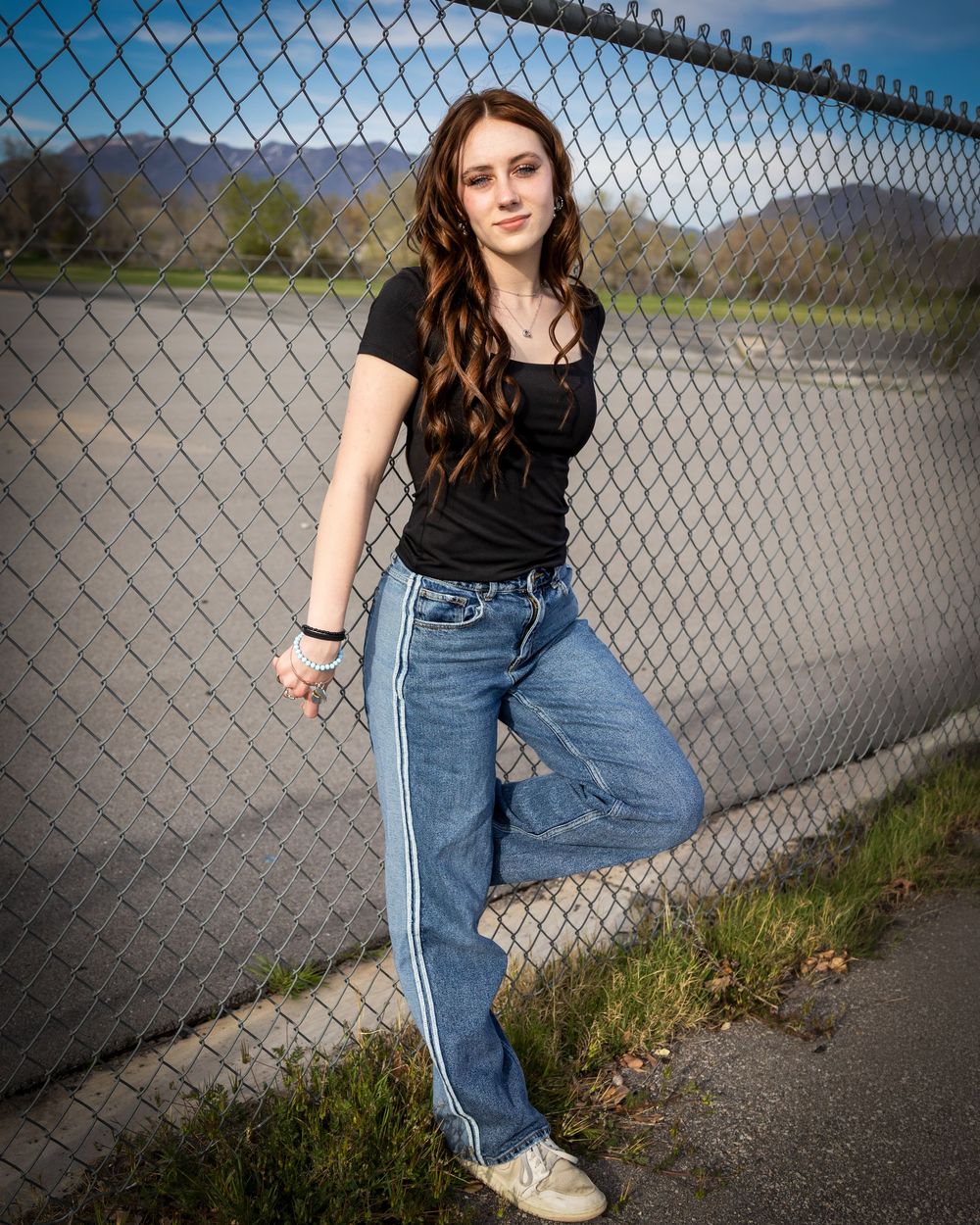 Young woman with long brown hair in black top and striped jeans leaning against chain-link fence with mountains behind.