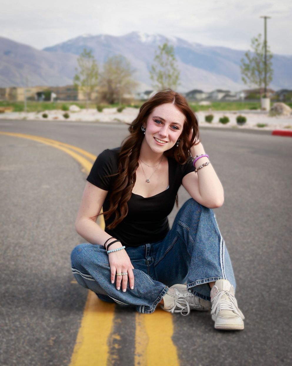 Young woman with red hair sitting on a road centerline, wearing black top and jeans, with mountains in the background.