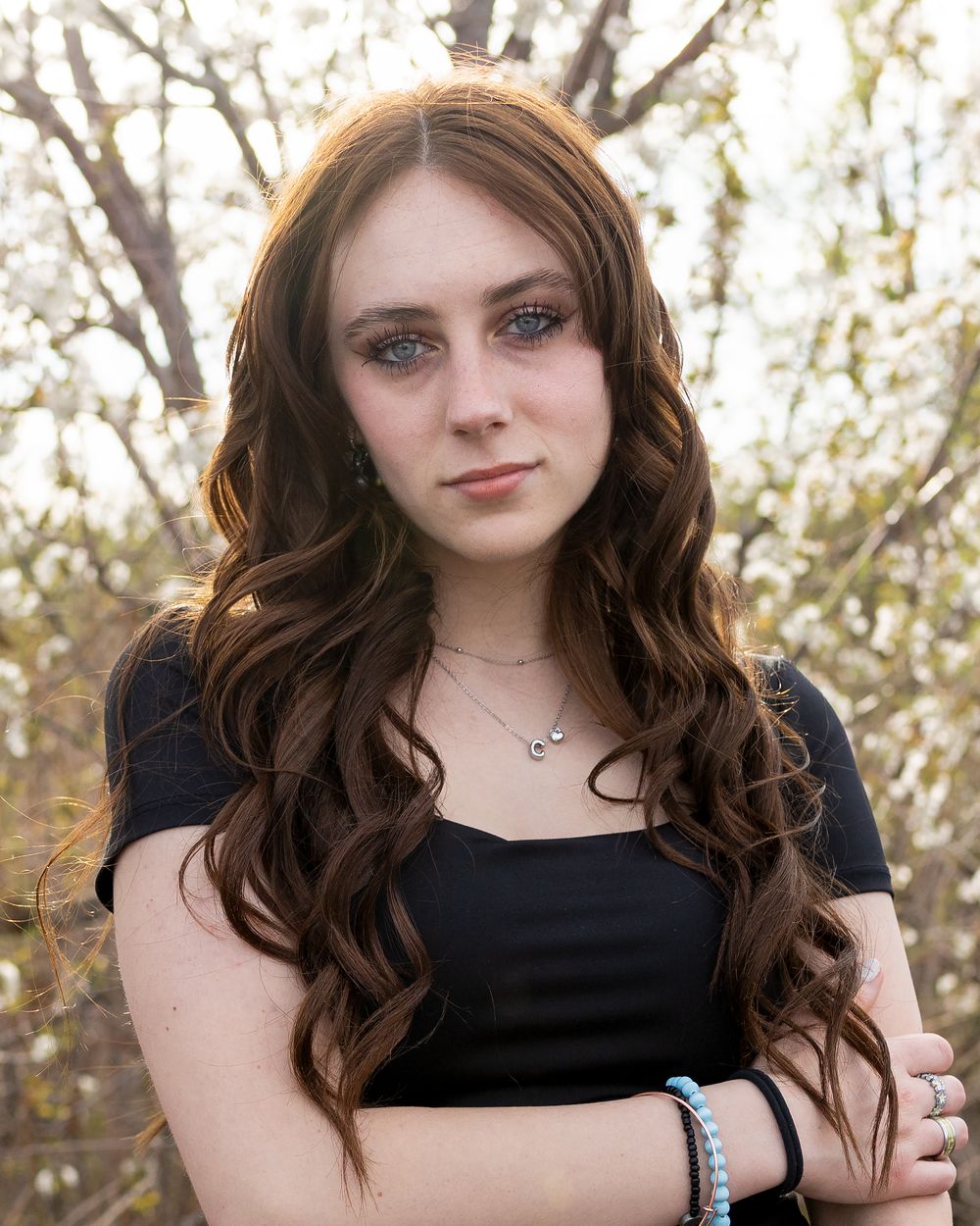 Young woman with long wavy auburn hair and blue eyes, wearing a black top, arms crossed, outdoors.