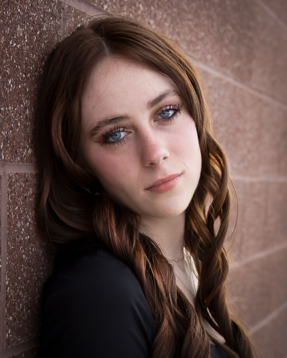 Young woman with blue eyes and auburn hair posing against a brick wall in a professional portrait photo.