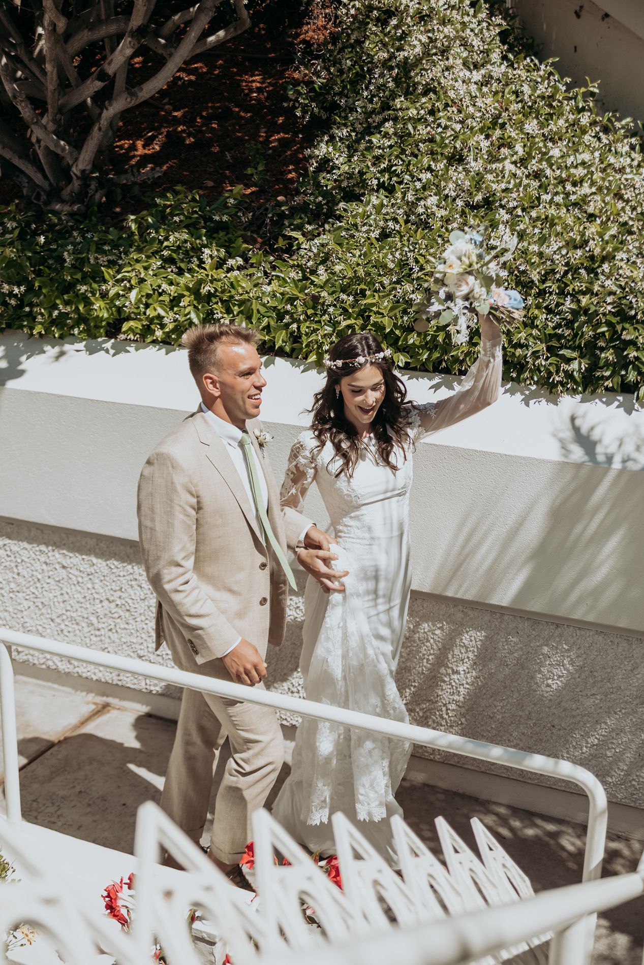 Bride and groom walking down stairs outdoors, bride raising bouquet, groom in tan suit, lush greenery background at LDS temple in San Diego.