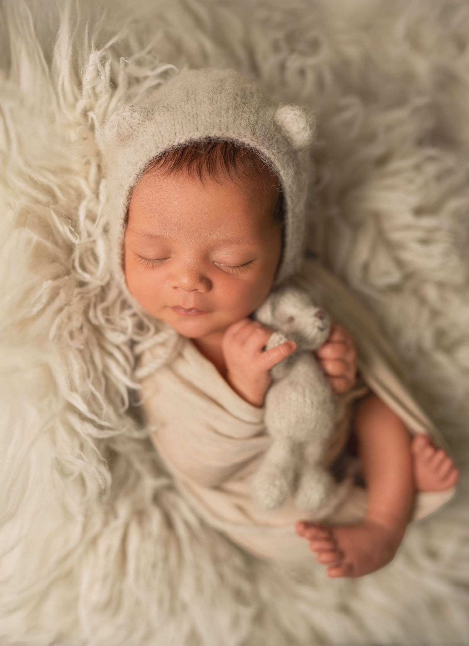 Newborn baby sleeping in a green wrap hammock, wearing a knit pom-pom bonnet, cradling a small brown teddy bear.