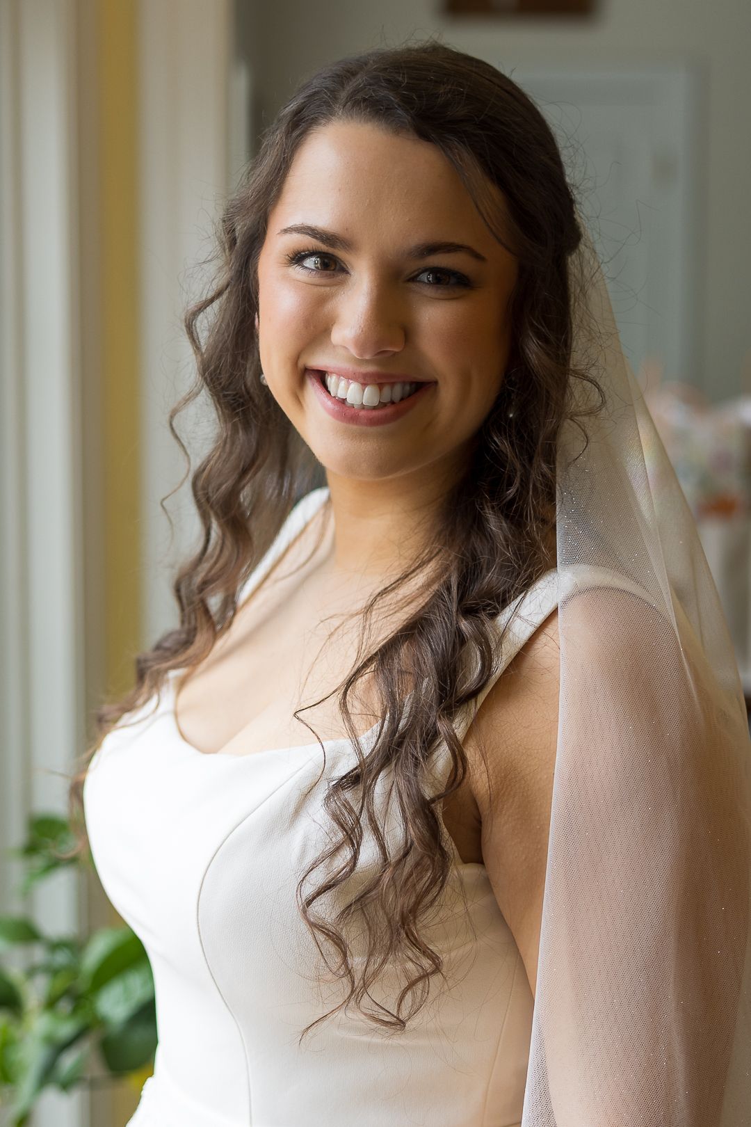 Smiling bride in white gown and veil with curly brown hair posing indoors near a window.