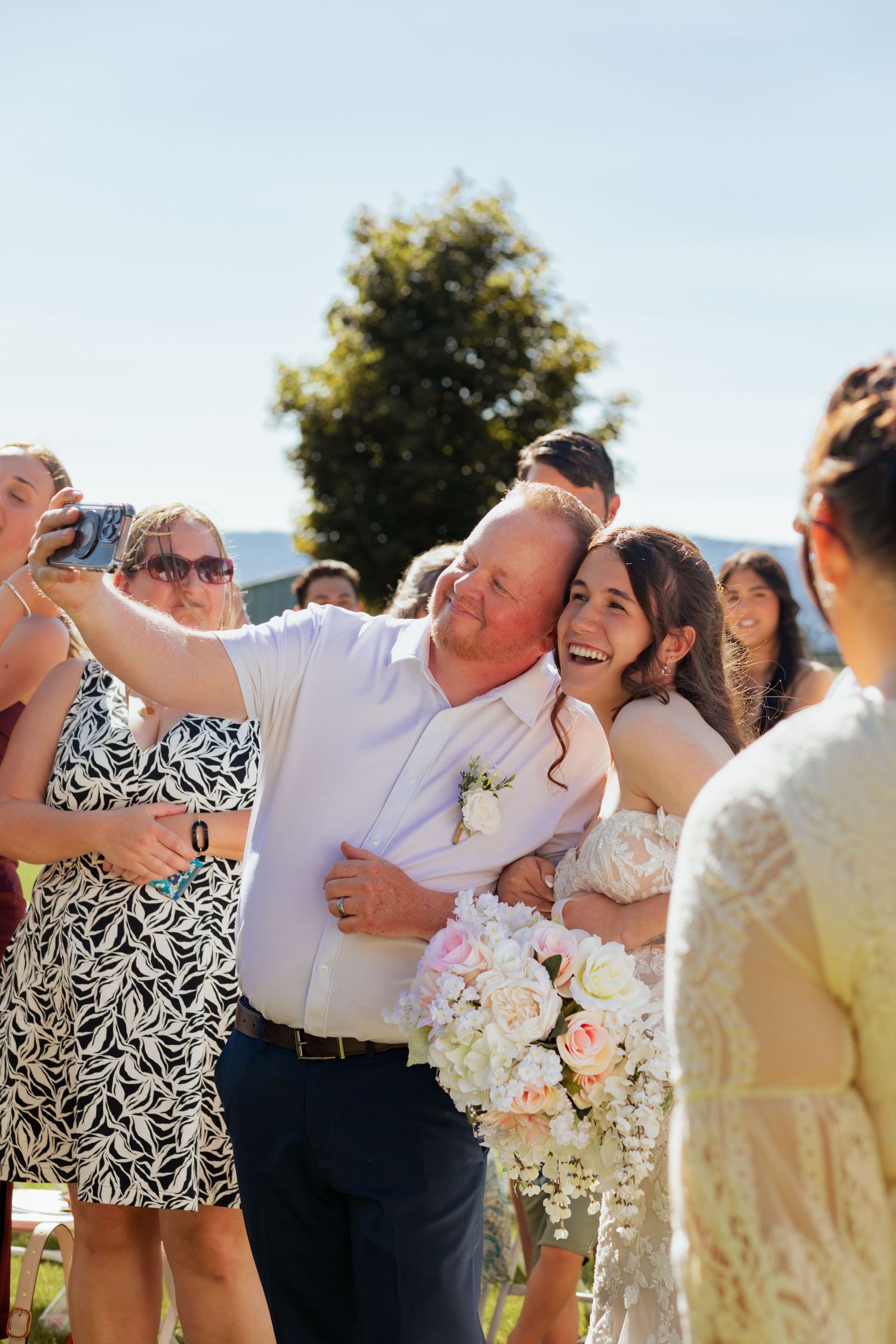 Bride laughing while taking a selfie with a guest at an outdoor wedding celebration on a sunny day.