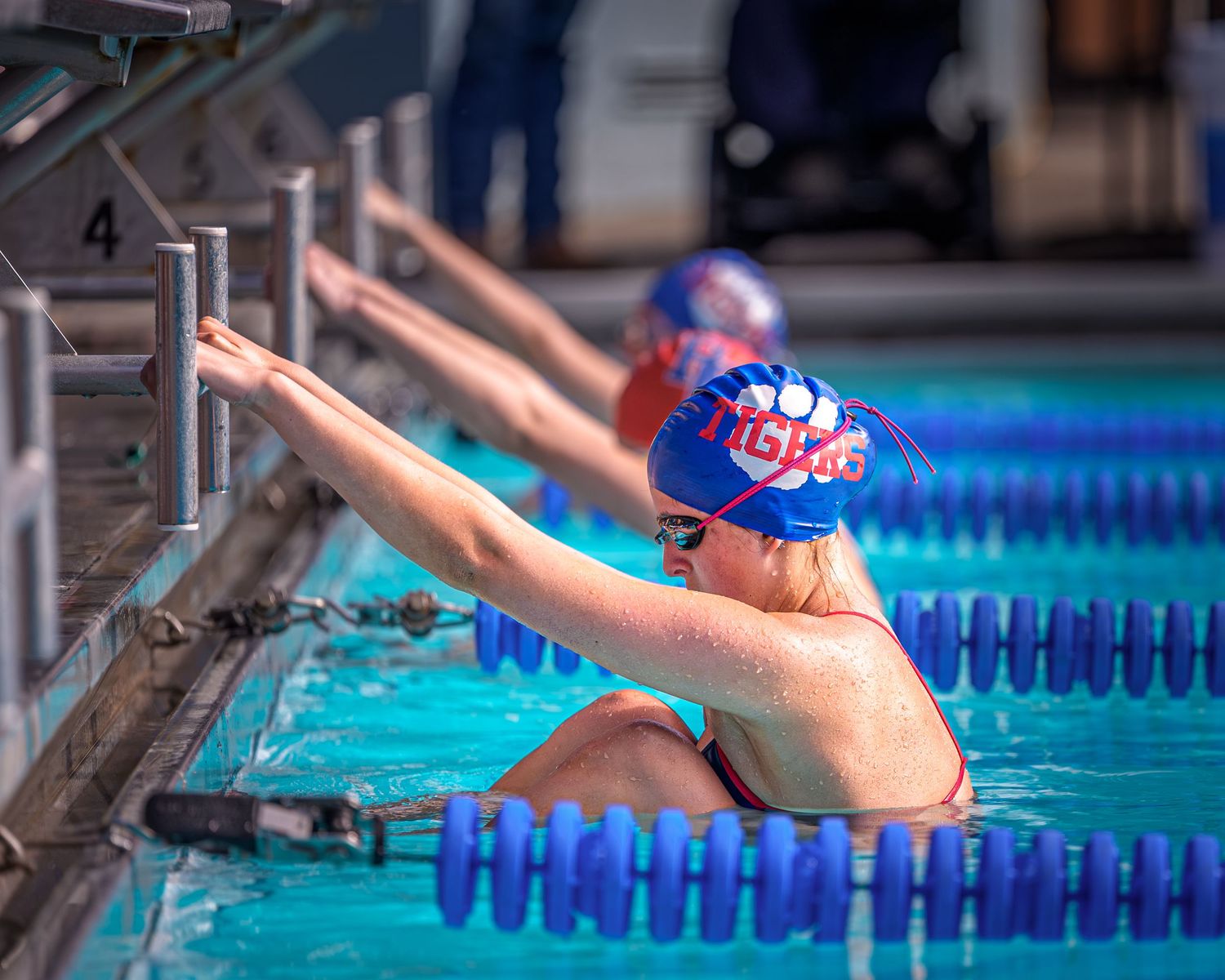 Competitive swimmer in a red cap grips the starting block, poised to begin a backstroke race in a blue lane pool.