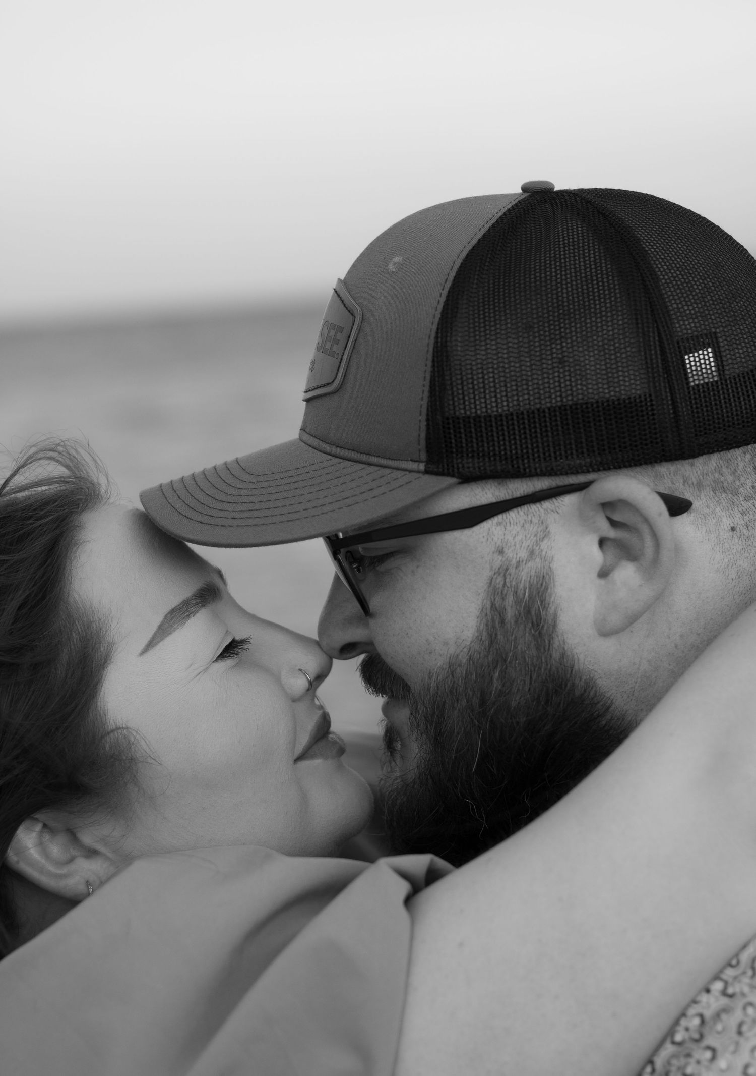 Black and white close-up of a couple in an affectionate embrace, nose to nose, with the man wearing a cap and glasses.