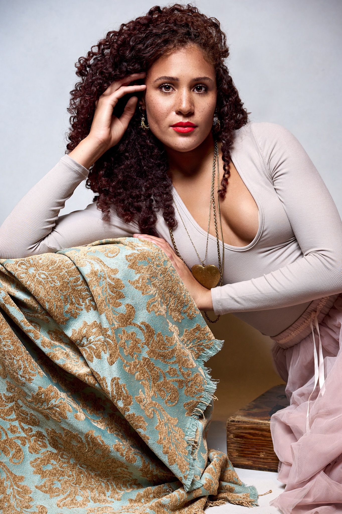 Woman with curly red hair in gray top posing with floral fabric in a soft studio setting.