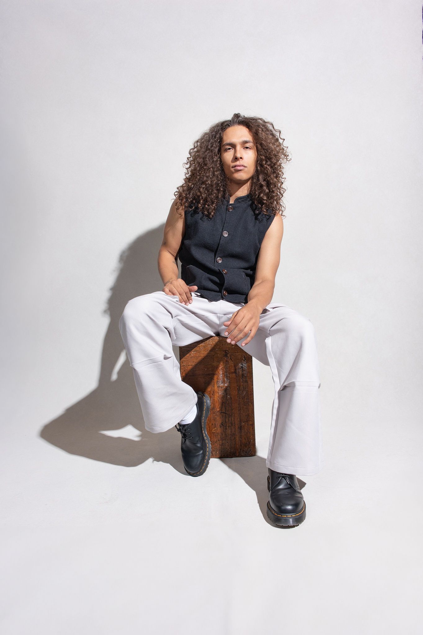Young man with long curly hair in black vest and white pants seated on a wooden box in studio.