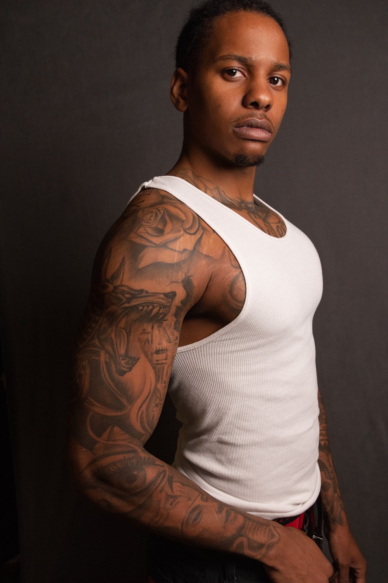Tattooed man in white tank top showcasing detailed sleeve tattoos against a dark gray background.