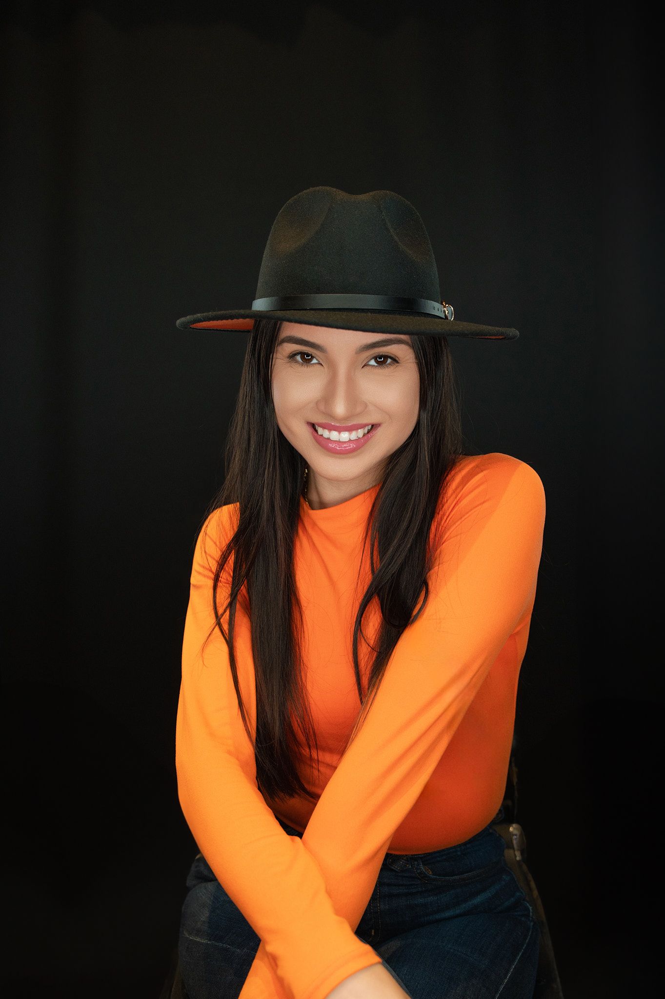 Smiling woman in orange top and wide-brim hat posing with crossed arms against a black background.