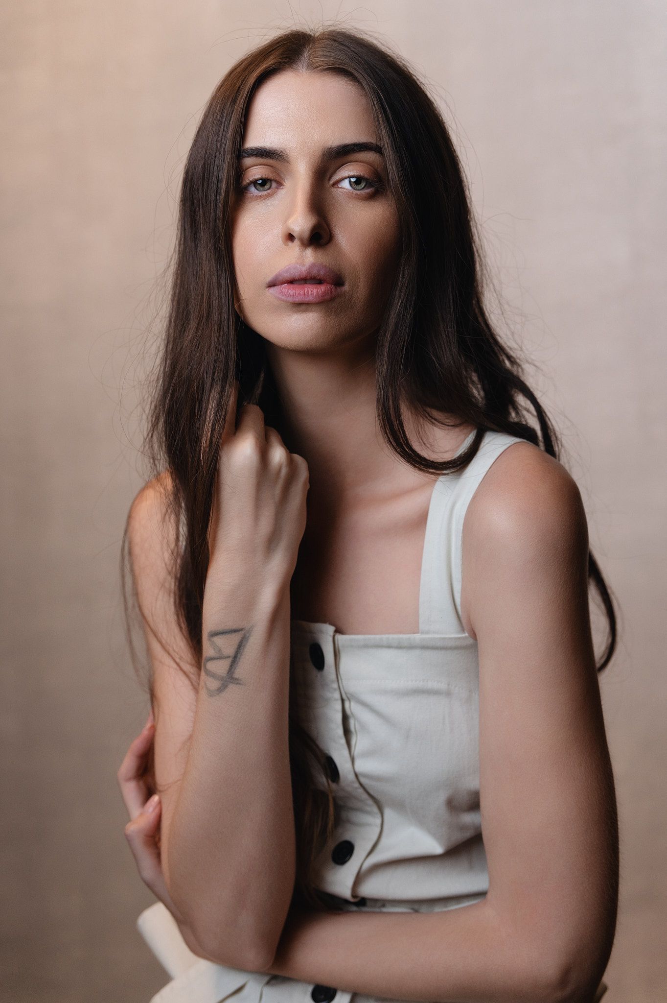 Young brunette woman with blue eyes poses in a white sleeveless top with a tattoo on her wrist.