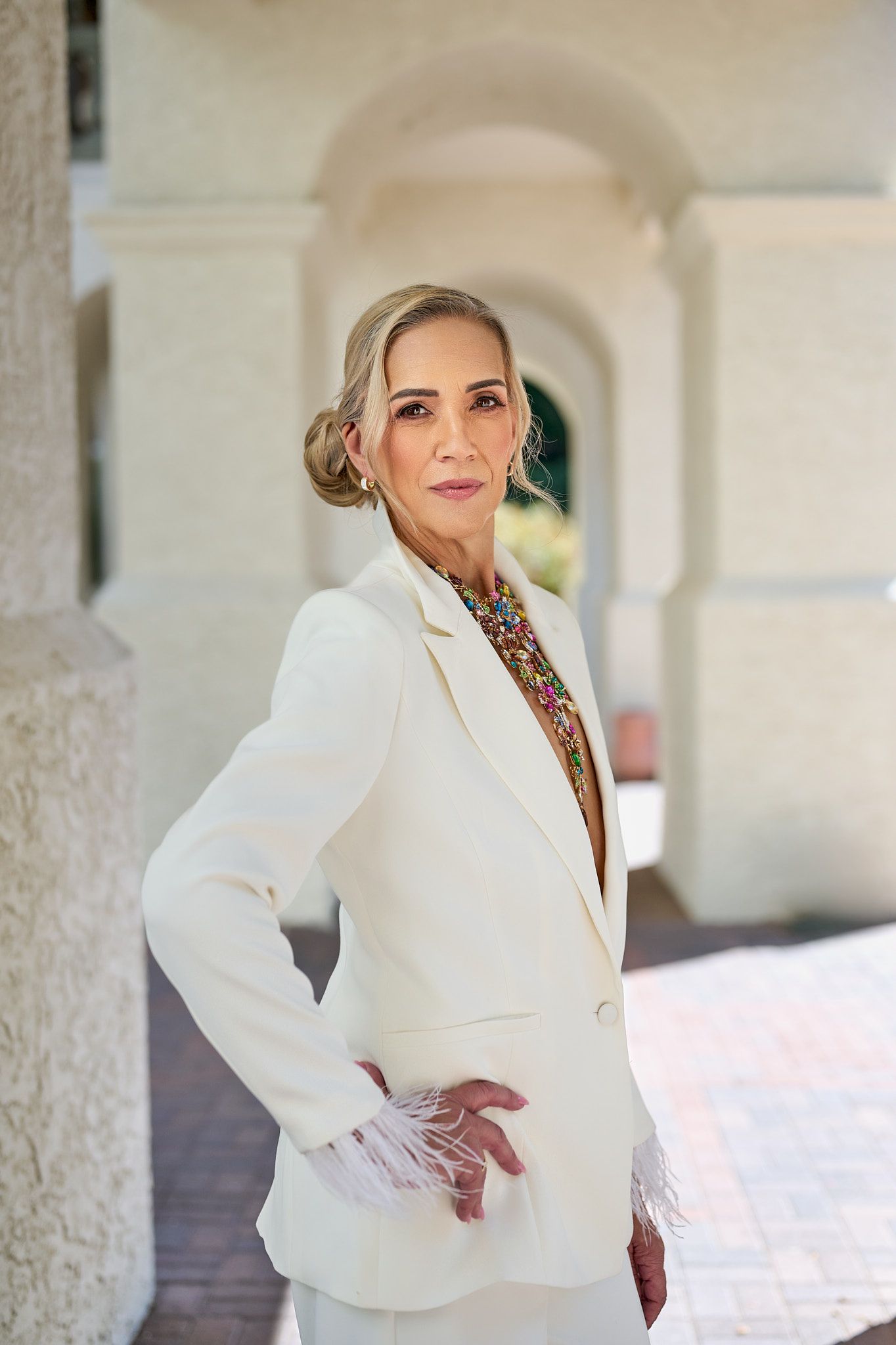 Elegant older woman in white blazer with feather trim posing confidently in an archway setting.