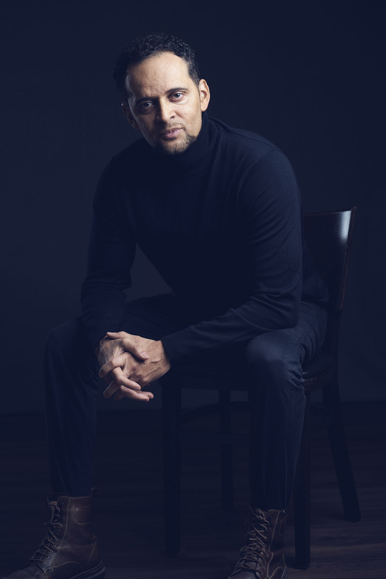 Man in black turtleneck and pants sitting in a dark studio, gazing intensely at the camera.