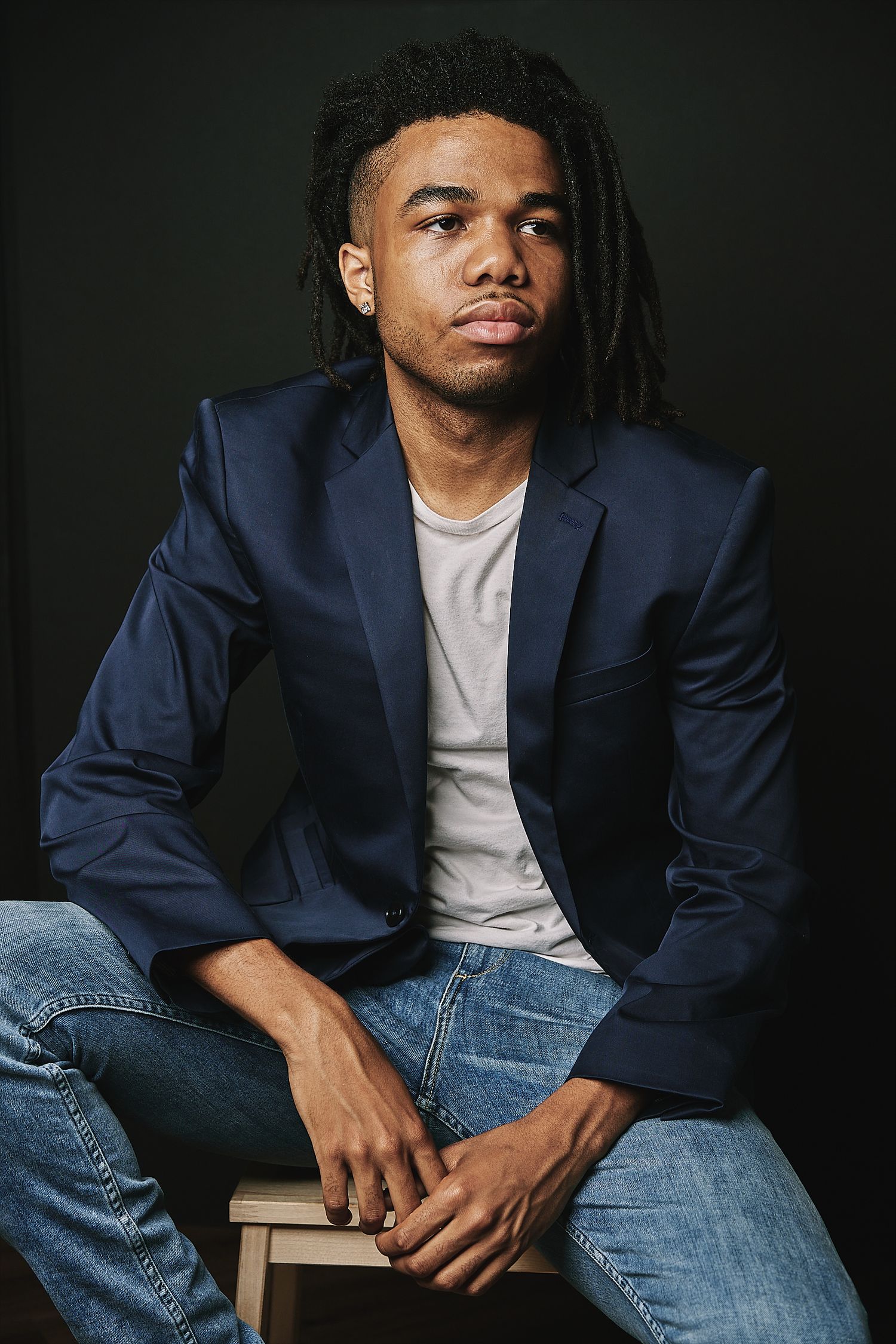 Stylish young man with dreadlocks wearing a navy blazer and jeans sits against a dark background.