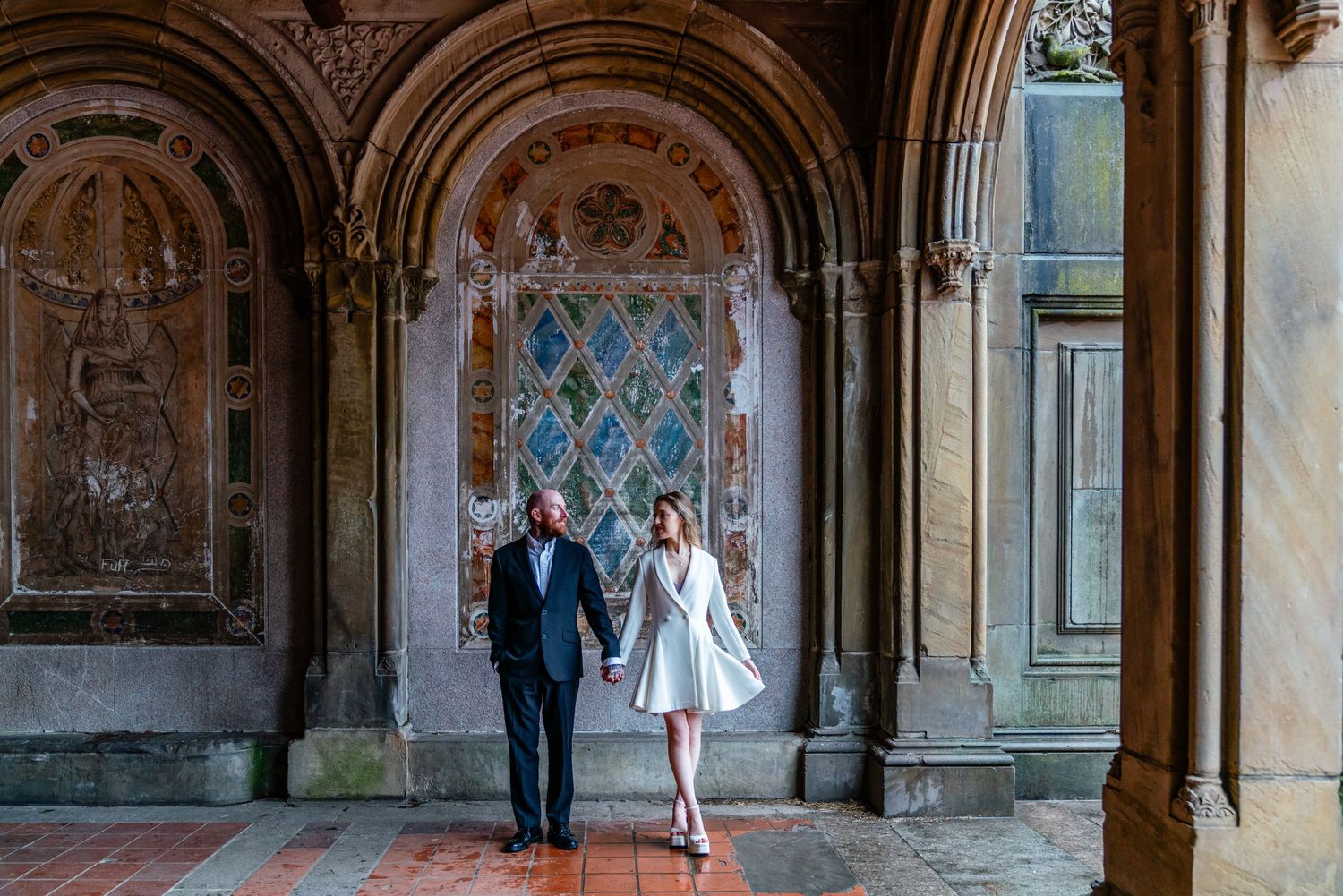 Couple holding hands at Bethesda Terrace in Central Park, NYC, with ornate arches and colorful tilework backdrop.