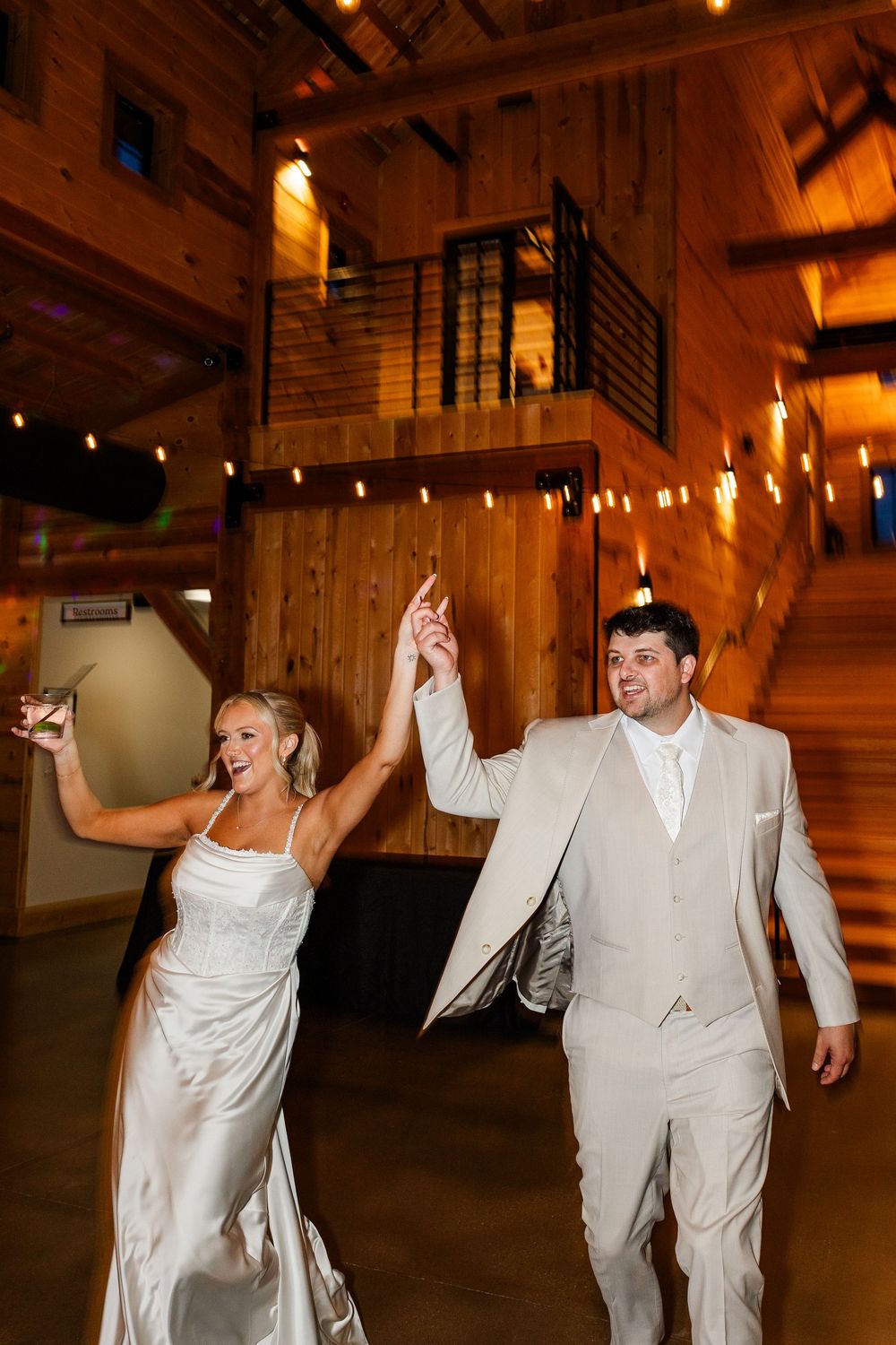 Bride and groom make grand entrance at rustic barn wedding reception, holding hands raised with warm string lights glowing.