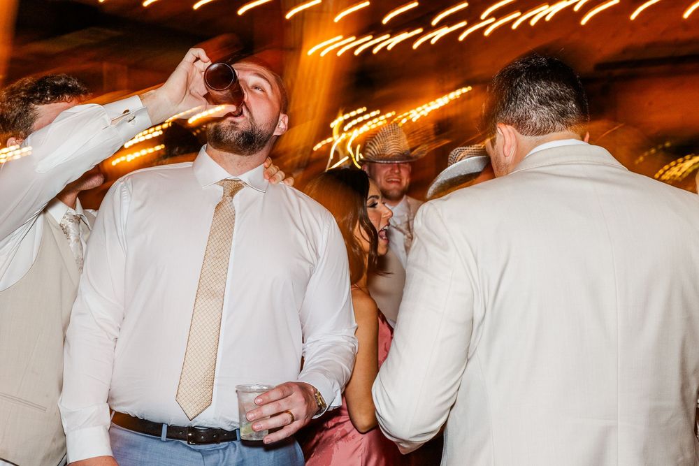 Men in dress shirts and ties celebrating at a wedding reception party, one drinking a shot glass on the dance floor.