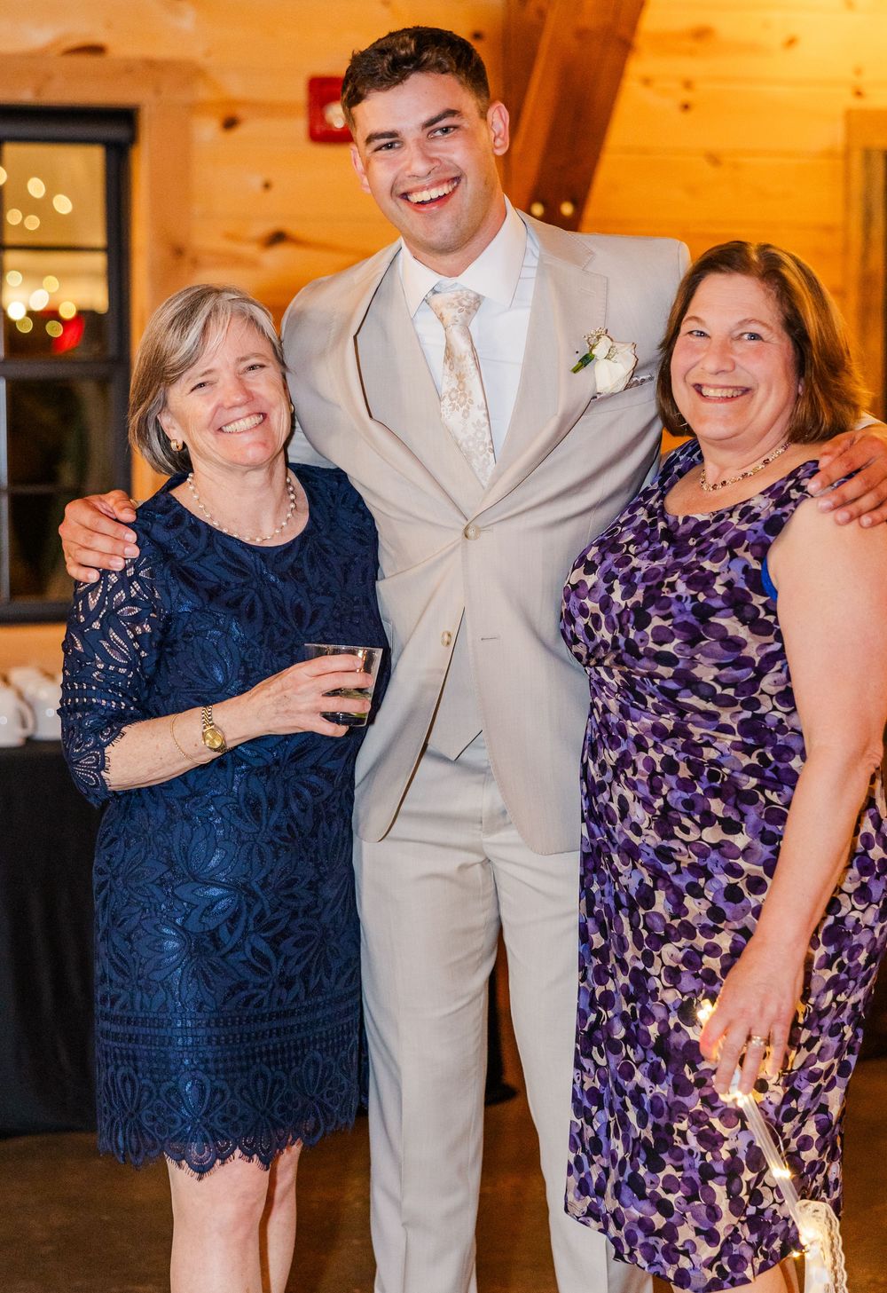 Groom in gray suit poses with two smiling women in formal dresses at an indoor wedding reception.