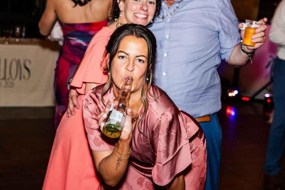 Woman in pink satin dress crouching on dance floor, playfully drinking from a glass at a lively party event.