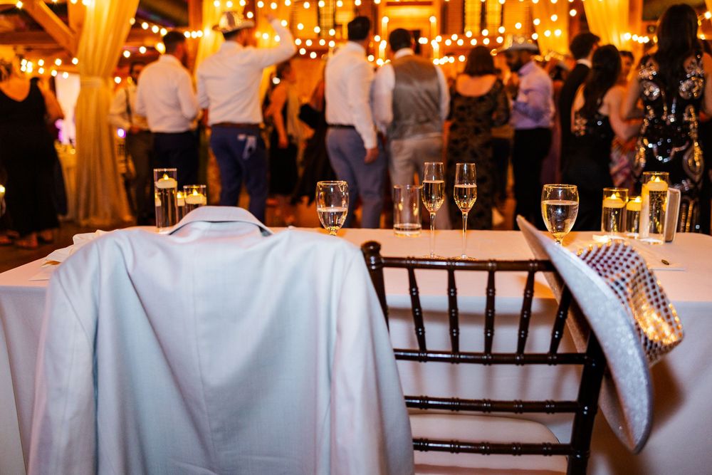 Wedding reception venue with champagne glasses, draped jacket on chair, string lights, and guests dancing in background.