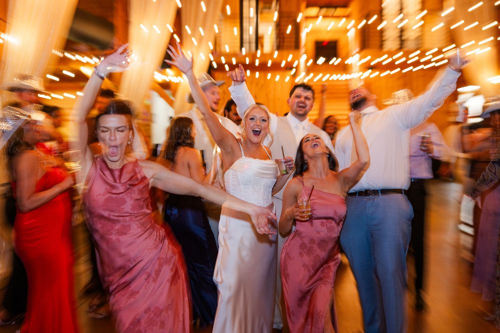 Bride and bridesmaids in pink dresses dancing joyfully at a wedding reception with warm string lights.