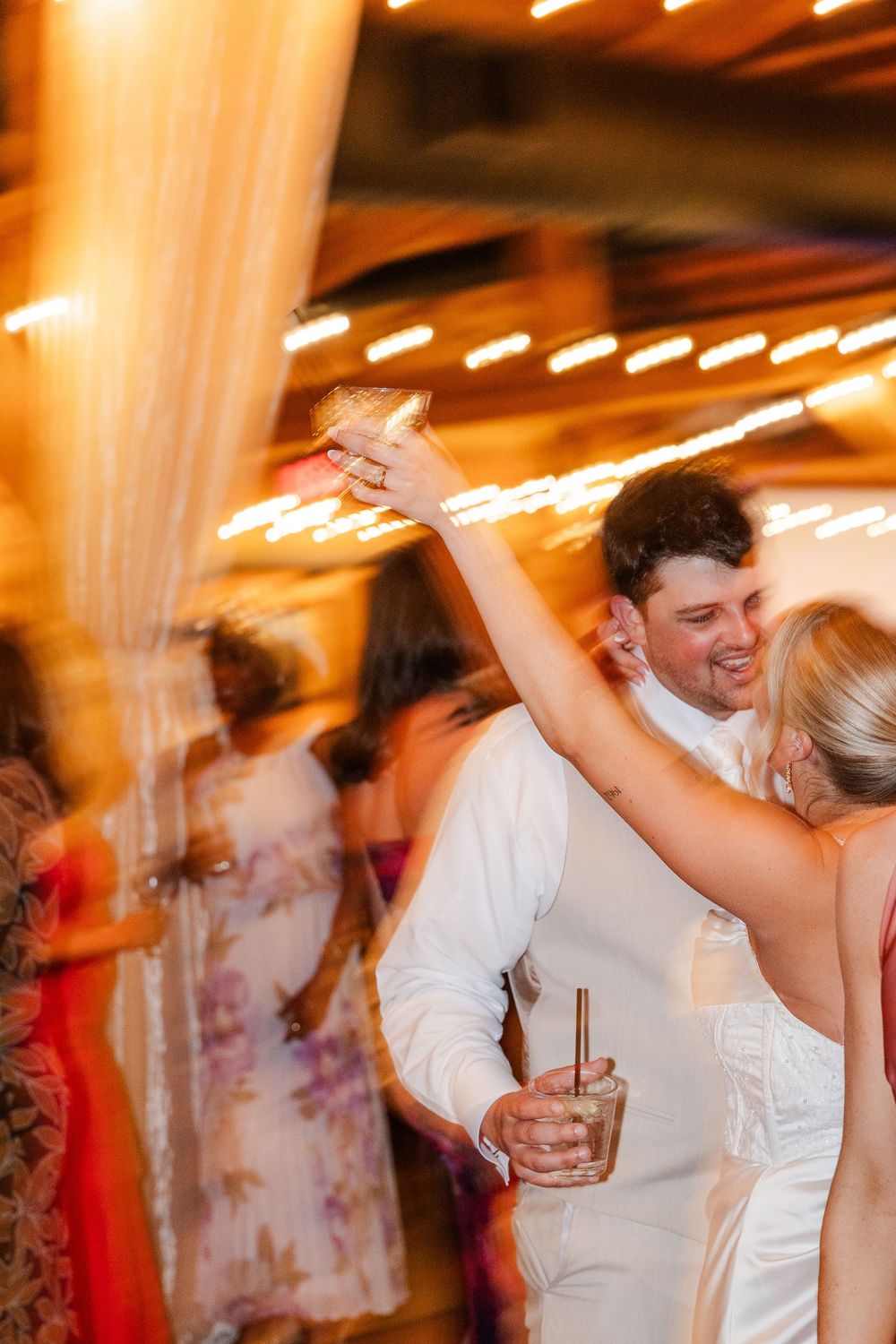 Bride and groom dancing joyfully at wedding reception, holding cocktails under warm string lights with guests in background.