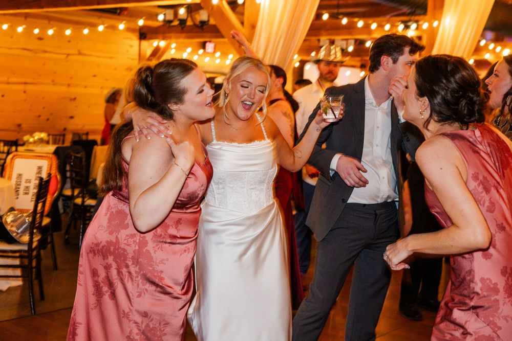 Bride in white gown dancing joyfully with bridesmaids in pink dresses at a warmly lit wedding reception.