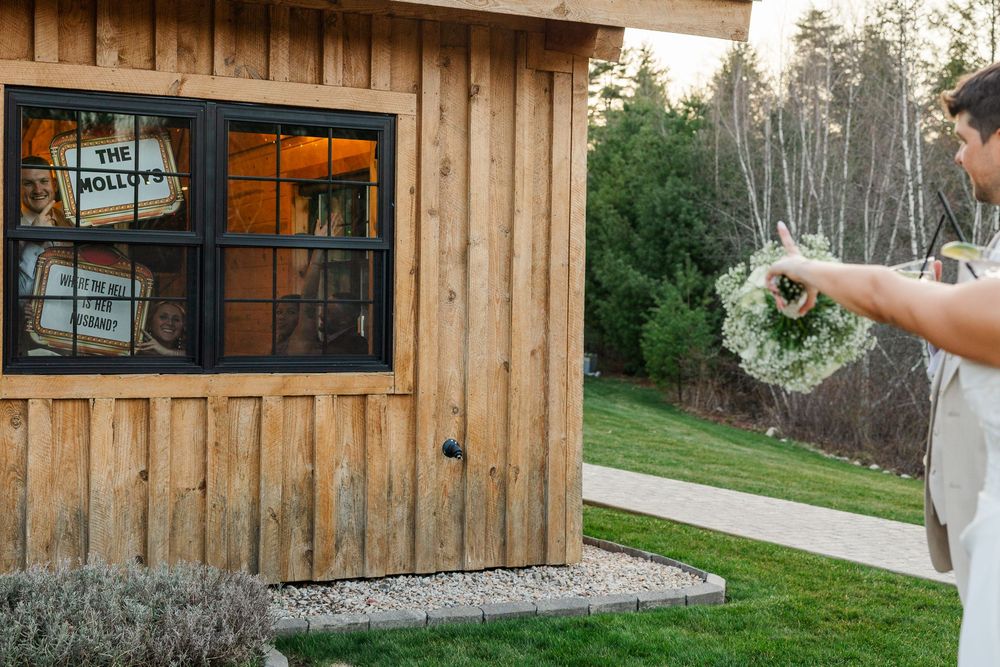 Man holding a floral wreath outside a rustic wooden cabin with black-framed windows in a lush garden setting.