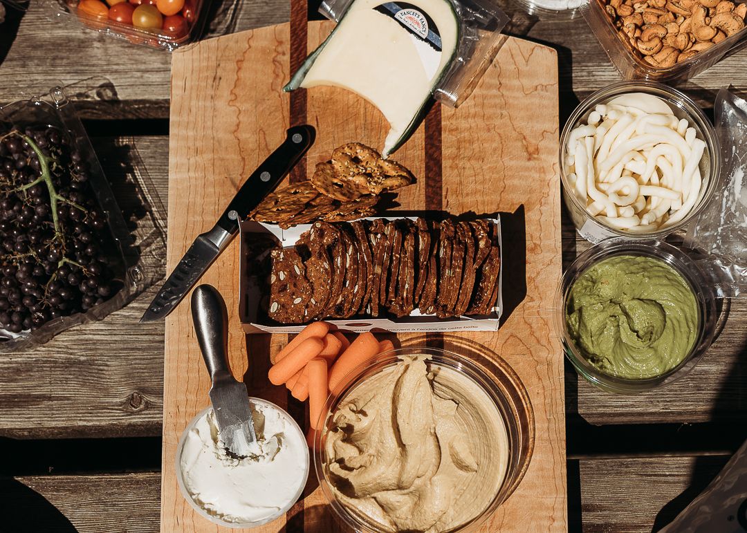Overhead view of a wooden charcuterie board with hummus, carrots, crackers, guacamole, and assorted dips outdoors.