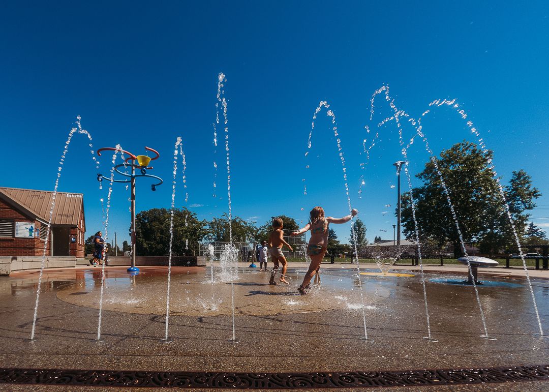 Children playing in outdoor splash pad fountains on a sunny day with clear blue sky and trees in background.