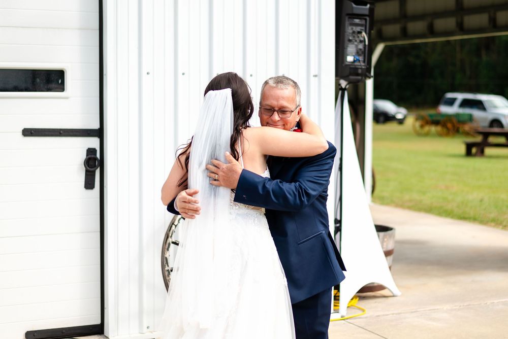 Bride and father share emotional first look hug outside white barn venue on wedding day.