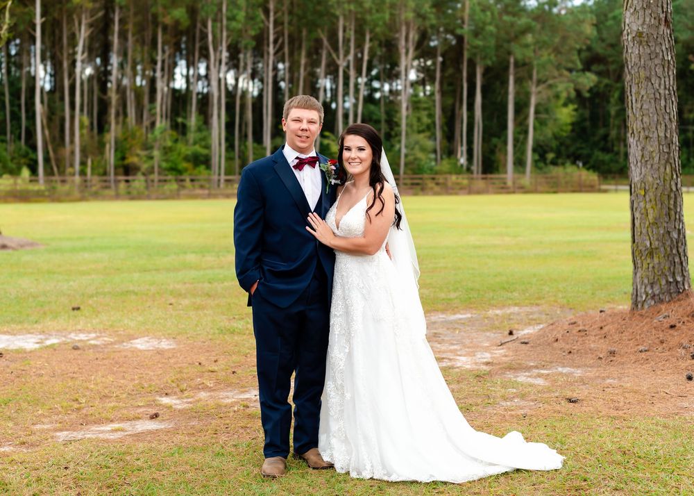 A bride in a white lace gown and groom in a navy suit pose together on a green lawn surrounded by pine trees.