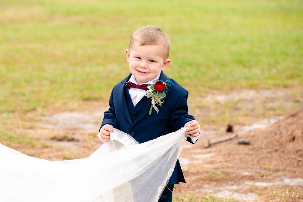 Cute ring bearer in navy suit and bow tie smiles while holding bride's wedding dress train outdoors.