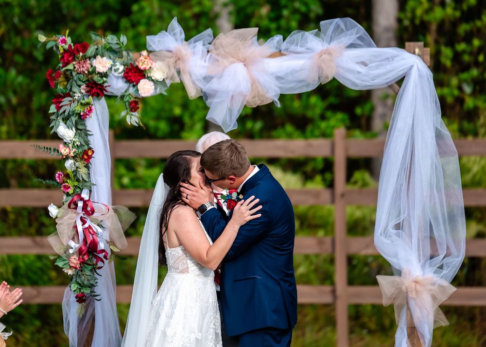 Bride and groom share first kiss at outdoor wedding altar decorated with red flowers and white tulle.