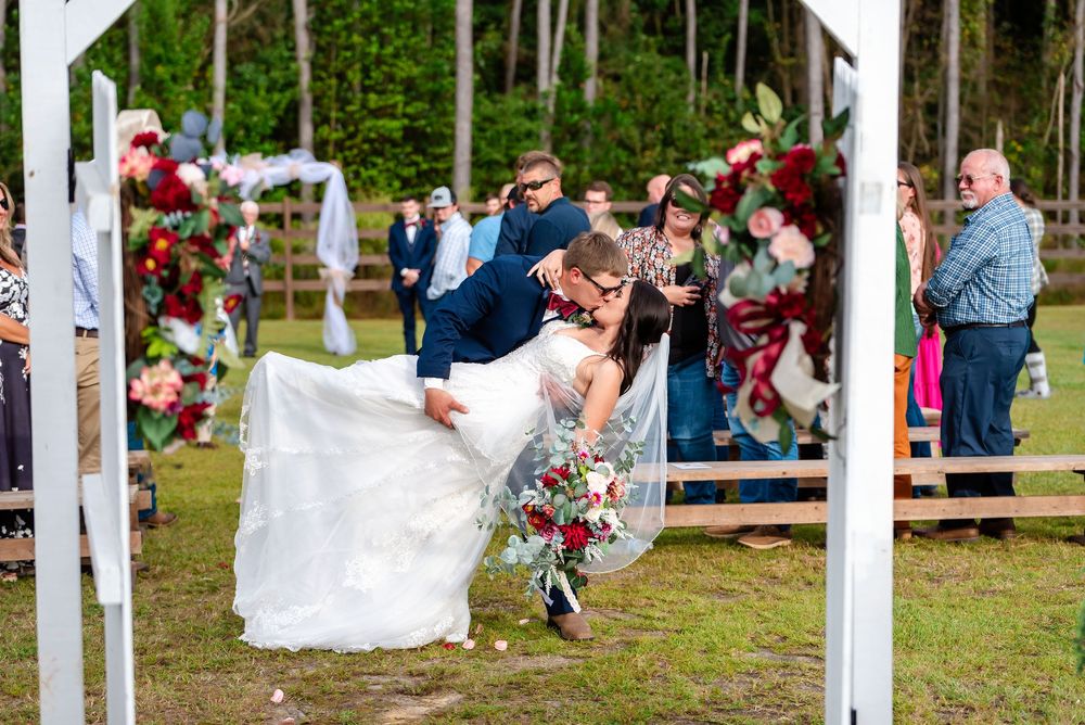 Groom dips bride for a kiss at outdoor wedding ceremony surrounded by floral arch and guests.